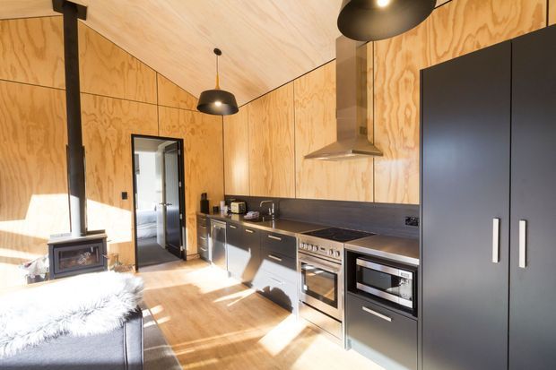 A compact open kitchen finished with warm plywood wall and ceiling panels, dark cabinetry, a dark benchtop and splashback, stainless-steel appliances, and black pendant lights. A freestanding wood-burning stove and chimney are visible at the left, with a doorway leading to another room. Sunlight creates strong highlights across the timber floor and cabinetry.