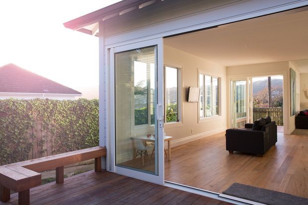 A bright contemporary open-plan living area viewed from a timber deck through wide-opening glazed doors. The room features warm timber flooring, light neutral walls, extensive windows, and a simple dark sofa, with views toward a leafy residential setting and hills beyond.
