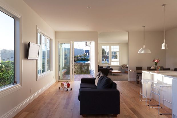 Bright open-plan living area with a dark sofa, timber flooring, large windows and glazed doors opening to a balcony or outdoor area. A white kitchen island with pendant lights and stools occupies the right side, while a dining or secondary seating area is visible beyond. The space has clean contemporary detailing and abundant natural light.