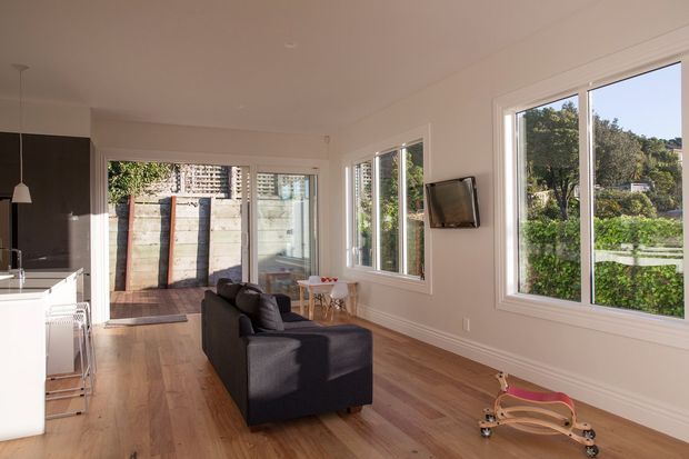 Bright open-plan living area with a compact white kitchen at left, dark sofa, timber flooring, white walls, and large windows overlooking greenery. Glazed doors open to a small timber deck and fenced outdoor area. A wall-mounted television and children's rocking toy are visible.