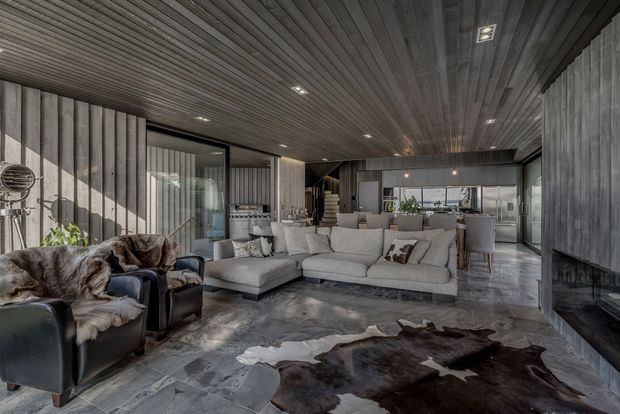 A spacious open-plan living room with a large light-grey sectional sofa, dark leather armchairs, a cowhide rug, and a dark stone-look floor. The room features extensive vertical and horizontal dark timber lining on the walls and ceiling, recessed lighting, large glazed openings, and a contemporary kitchen visible at the rear. A substantial dark fireplace or hearth feature occupies the right side.