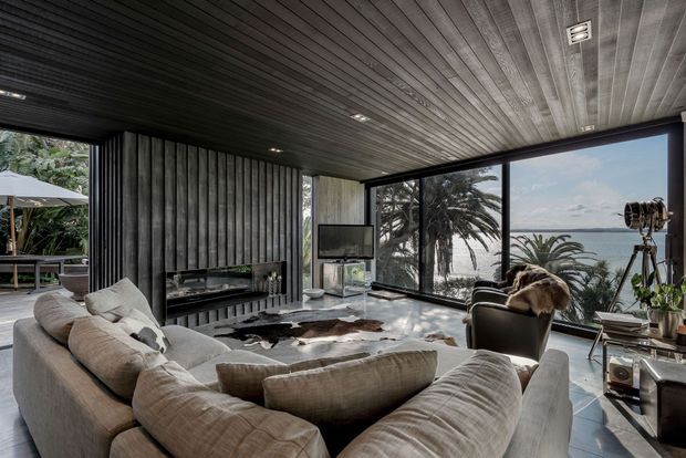 A dramatic contemporary living room with a large curved sectional sofa facing a low entertainment unit and television. The room features dark vertically ribbed wall panelling, matching linear ceiling boards, expansive corner glazing, a sculptural black lounge chair, a cowhide rug, and an industrial-style spotlight. Lush tropical vegetation and a bright coastal-looking landscape are visible beyond the glass.
