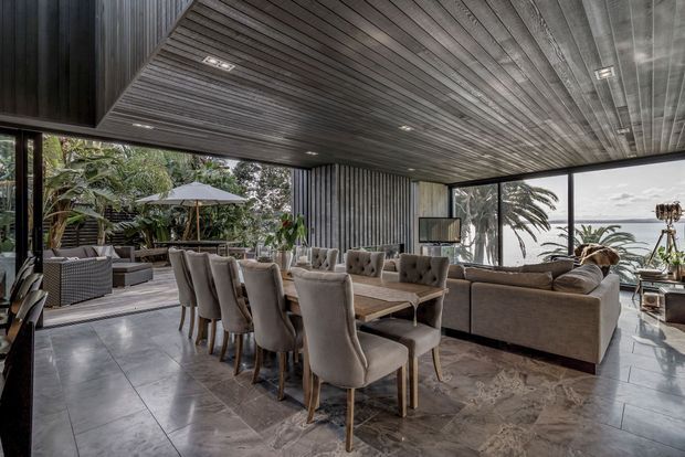 A sophisticated open-plan living and dining area with a long upholstered dining table and chairs in the foreground, and a large sectional sofa arranged around a television in the lounge area. Dark vertically ribbed wall cladding and a dramatic slatted timber ceiling frame expansive glazing, which opens the room visually to a lush tropical garden and coastal view. The space has large-format grey stone-look floor tiles, subdued neutral furnishings, and integrated recessed lighting.