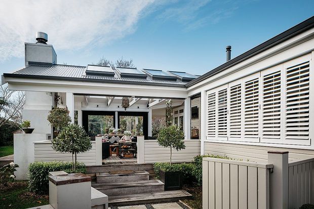 Exterior view of a low-rise home with a covered outdoor entertaining area, broad veranda, timber privacy shutters, light-coloured weatherboard walls, and a corrugated metal roof with multiple skylights. Steps lead through landscaped planting to the raised outdoor space, with a partially visible outdoor lounge and dining area beyond.