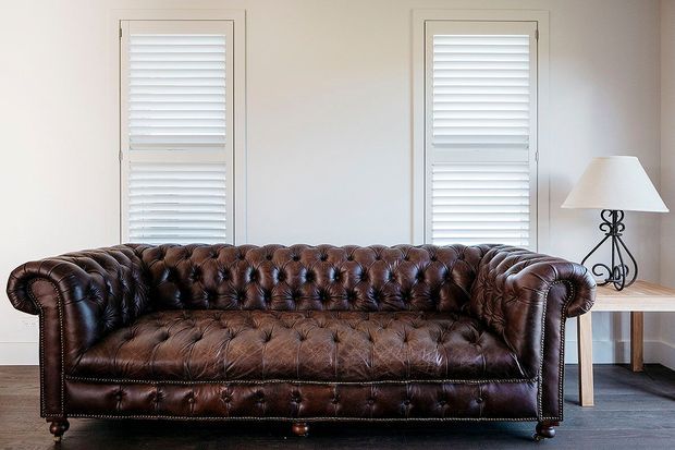 A traditional living room featuring a large deep-buttoned brown leather Chesterfield sofa with rolled arms and nailhead trim. Two tall windows with white plantation shutters sit behind the sofa, while a small light-wood side table and decorative table lamp occupy the right side. The room has pale walls and dark flooring, creating a classic, refined interior.