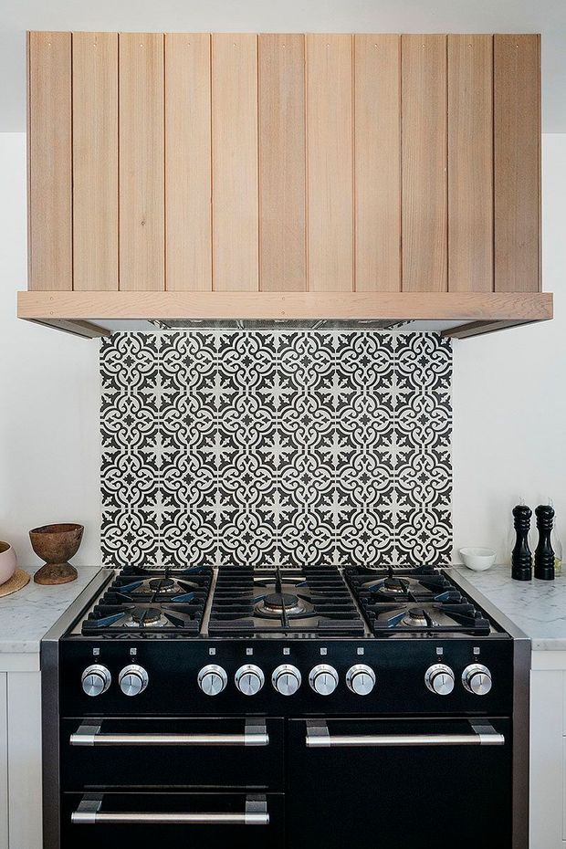 A close-up view of a traditional-style kitchen cooking area featuring a large black freestanding range cooker with multiple gas burners and stainless-steel handles. Above it is a pale timber rangehood with vertical slatted cladding, while the backsplash is covered in a bold black-and-white patterned tile. White cabinetry and pale stone-look benchtops frame the cooking zone, with pepper mills and small decorative vessels on the counters.
