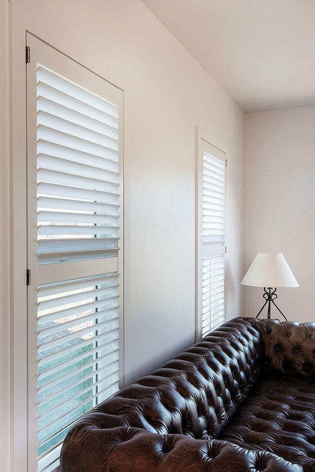 Bright, compact living room corner with two tall windows fitted with white plantation shutters. A dark brown tufted leather Chesterfield-style sofa occupies the foreground, paired with a small table lamp with a white shade. The walls, ceiling, window surrounds and shutters are predominantly white, creating a clean, traditional interior with strong natural light.