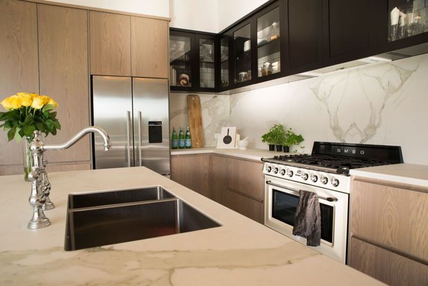 A refined contemporary kitchen with a large pale stone island and integrated double sink, stainless-steel French-door refrigerator, freestanding stainless-steel range, pale timber-grain cabinetry, dark upper cabinets with glass-front display doors, and a dramatic white marble-look splashback. Warm pendant-free task lighting and small potted greenery add a welcoming residential feel.