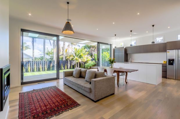 Bright open-plan living, dining and kitchen interior with expansive floor-to-ceiling glazing opening toward a leafy outdoor area. The room features pale timber flooring, white walls and cabinetry, a grey upholstered sofa, a timber dining table, a patterned area rug, dark-framed sliding doors, and a stainless-steel refrigerator. Recessed ceiling lights and pendant fixtures provide contemporary illumination.