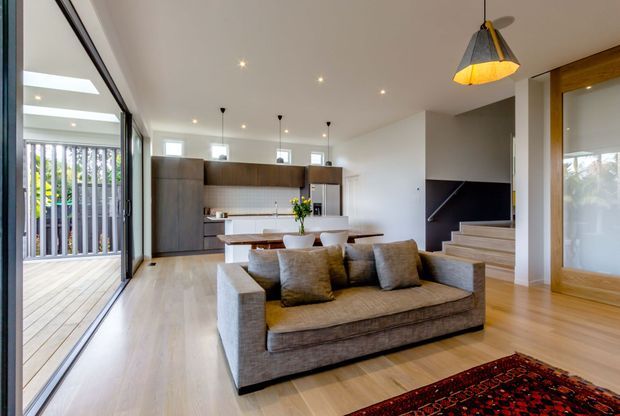 Open-plan contemporary living area with a large upholstered sofa facing a compact kitchen and dining zone. The room has pale timber flooring, white walls and ceiling, extensive floor-to-ceiling glazing opening to a timber deck, dark wood kitchen cabinetry, a timber-framed glazed wall, recessed downlights, pendant lighting, and stairs leading upward at the rear.