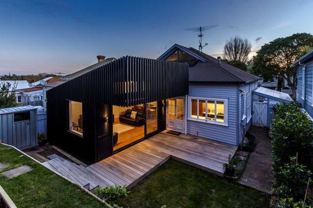 Twilight exterior view of a compact residential extension combining a dark vertically slatted pavilion with an existing light-grey weatherboard house. The addition has large glazed openings, warm interior lighting, and opens onto a timber deck and small landscaped yard. The original house has a pitched metal roof and gabled upper section, with neighbouring homes visible in the background.