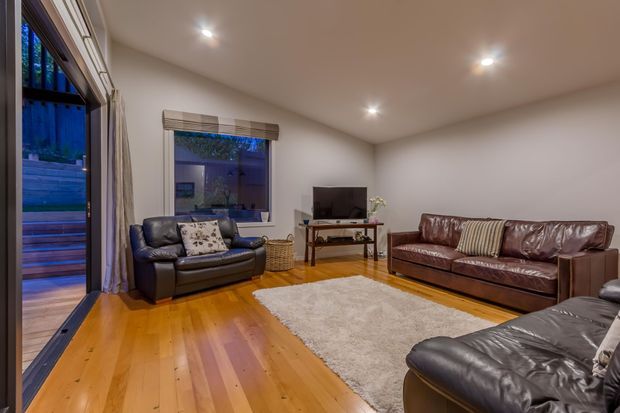 A spacious living room with polished warm timber flooring, cream walls, recessed ceiling lights, and large glazed openings with curtains. The room contains dark brown leather sofas and armchairs, a television on a simple timber console, a pale area rug, and a woven basket. Outdoor greenery and decking are visible through the glazing.