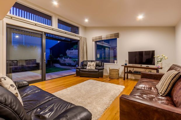A warm, contemporary living room with polished timber flooring, leather sofas and armchairs, a large light rug, recessed ceiling lights, and a television on a low console. Full-height sliding glass doors and upper clerestory-style glazing open toward an outdoor garden area, while patterned curtains and cushions add modest decorative detail.