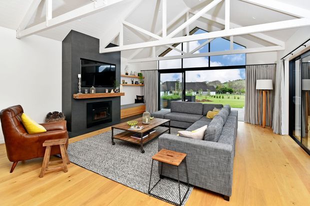 Bright, open-plan living room with a large grey sectional sofa, leather armchair, timber coffee table, black fireplace surround, and wall-mounted television. White exposed roof trusses and beams create a high pitched ceiling, while full-height glazing opens to a green landscape. Light timber flooring and neutral furnishings give the space a warm, contemporary feel.
