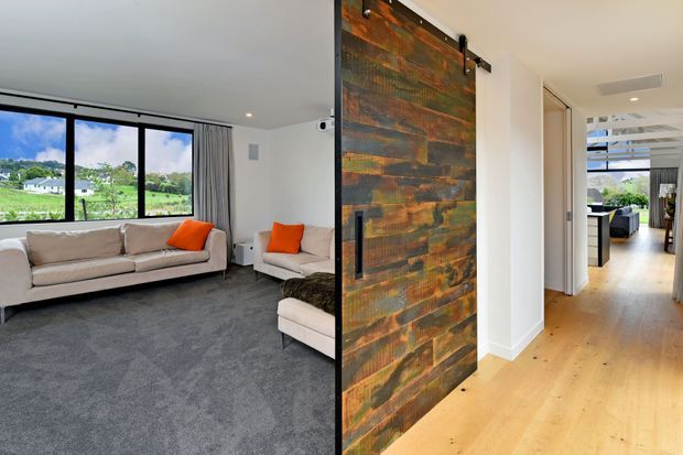 A bright contemporary living room opening to a hallway, with large black-framed windows overlooking greenery, pale upholstered sofas with vivid orange cushions, and a dark grey carpet. A striking sliding barn-style door clad in weathered multicoloured timber forms a feature between the living area and the light timber-floored hall. Additional living and dining areas are visible deeper in the home.