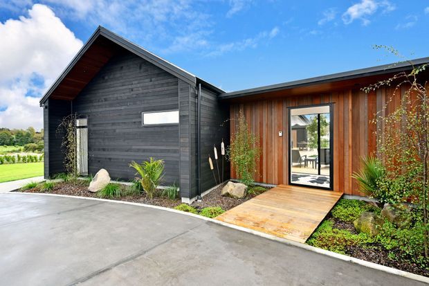Exterior view of a contemporary single-storey building with contrasting dark timber-clad gabled and warm vertical timber-clad sections. A glazed entry door opens onto a small timber deck and landscaped garden, with a concrete driveway in the foreground and rural greenery beyond.