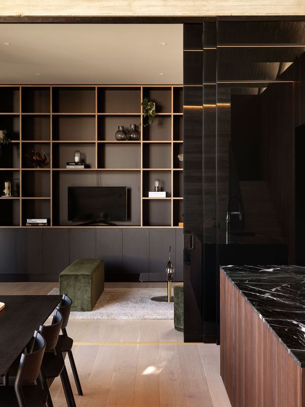 A dark, refined open-plan interior showing a living room and dining area beside a kitchen. The living room features a full-width built-in shelving and media wall with dark cabinetry and warm timber grid framing, a television, decorative objects, a green upholstered ottoman, and a light rug. In the foreground is a dark dining table with leather chairs, while the right side shows a kitchen island or benchtop with a dramatic black-and-white veined stone surface and dark timber cabinetry. Warm timber flooring, black finishes, and integrated lighting create a sophisticated, moody atmosphere.