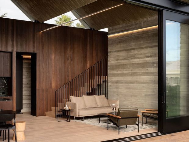 A refined open-plan living space with a warm timber ceiling and flooring, dark timber wall panelling, a textured concrete feature wall, and expansive black-framed sliding glass doors opening to the outdoors. A beige sofa and mid-century-style lounge chairs are arranged around a low coffee table, with a visible stairway rising behind the seating. Linear integrated lighting emphasizes the ceiling and wall junctions.