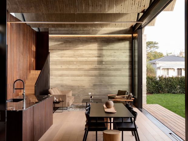A refined open-plan living, dining and kitchen interior with a warm timber-lined ceiling and kitchen cabinetry, a dark stone island benchtop, and a horizontal timber feature wall. Black-framed full-height glazing opens directly onto a timber deck and lawn, bringing in abundant natural light. The space is furnished with a dark dining table and chairs, a low coffee table, lounge seating and a timber-framed armchair, creating a sophisticated indoor-outdoor connection.