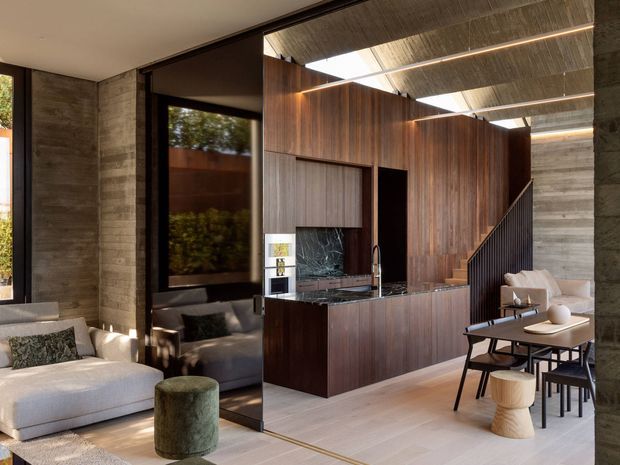 A sophisticated open-plan living and dining area with a double-height timber-lined volume, integrated dark timber kitchen or media joinery, exposed concrete walls, and a dramatic skylit ceiling. The space includes a light upholstered sofa, lounge chair, compact dining table, and a stair with dark balustrade. Floor-to-ceiling glazing and a large reflective dark panel bring in natural light and greenery.