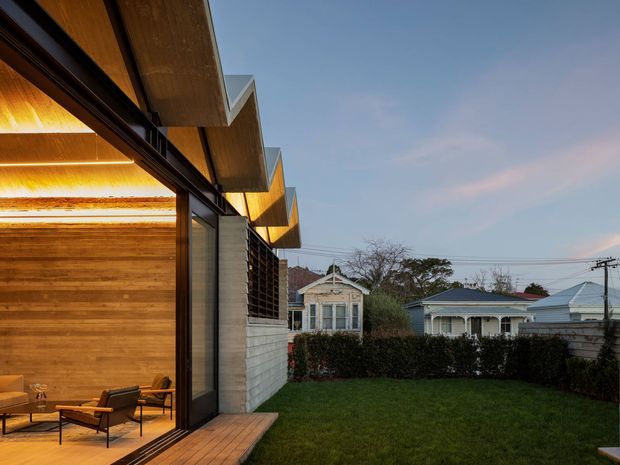 Exterior architectural view of a contemporary home extension opening onto a lawn and suburban garden. The building features extensive floor-to-ceiling glazing, warm timber wall and soffit lining, dark slender window framing, projecting angular roof forms, and a concrete block side wall. A furnished living area is visible through the open glazing, while neighbouring residential houses appear beyond the hedge.