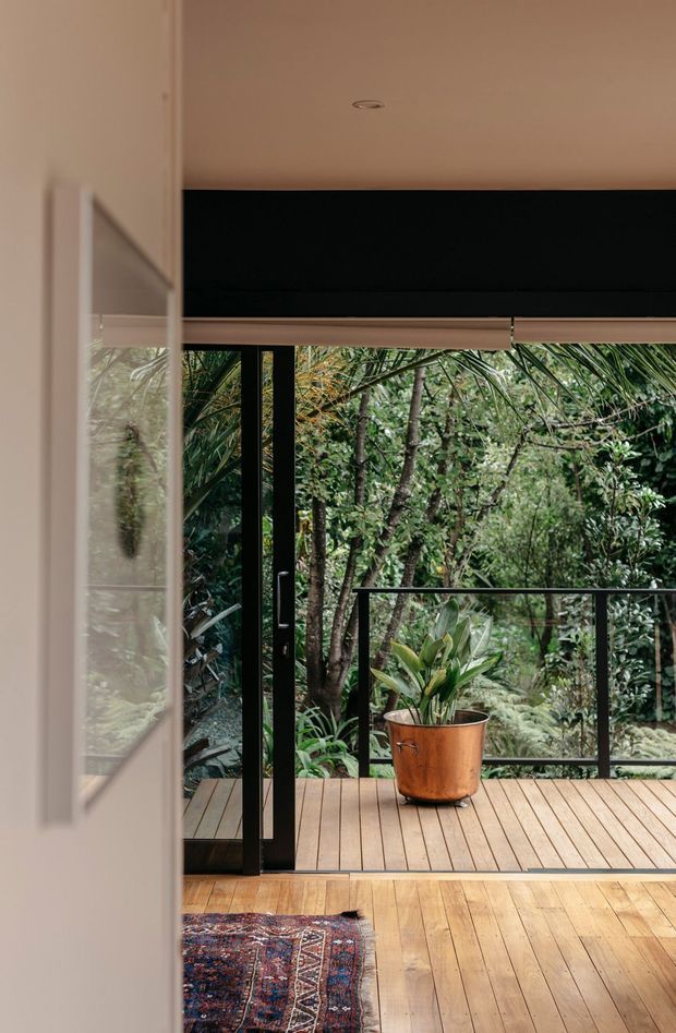 Interior view looking through a glazed opening onto a timber deck surrounded by dense greenery. The space features warm timber flooring, a partially visible patterned rug, black-framed sliding or folding glazing, a black roller blind or overhead blind housing, and a copper-toned planter. The image has a calm, nature-connected indoor–outdoor character.