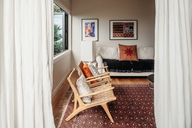 A cozy, compact living room viewed through sheer white curtains. The room features a cream sofa with a dark fringed throw, a pair of woven timber lounge chairs with cushions, a patterned red and dark rug, framed artwork, timber flooring, and a large opening window overlooking greenery.
