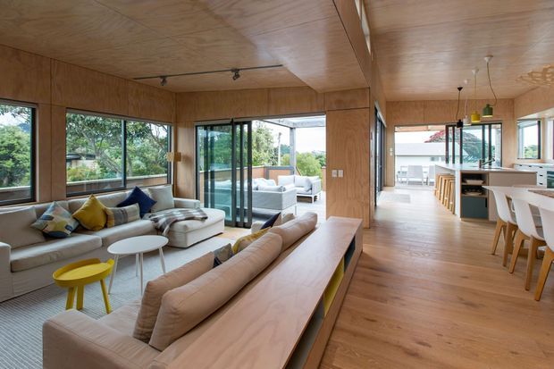Open-plan contemporary living and dining area clad in warm natural timber, with large windows and sliding glass doors connecting to an outdoor terrace. A sectional sofa, built-in timber-backed seating, coffee tables, and a bright dining/kitchen zone create an airy, connected interior.