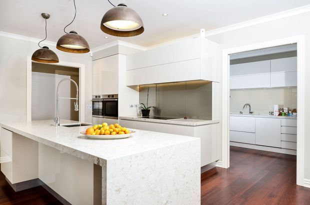 Bright contemporary white kitchen with a large waterfall-edge stone island, integrated white cabinetry, stainless-steel appliances, a metallic splashback, timber flooring, and three dark pendant lights. A second galley-style kitchen or utility area is visible through the wide opening on the right.