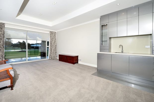 Bright open-plan living area with a contemporary built-in kitchen along the right wall, featuring sleek grey cabinetry, a pale backsplash, integrated appliances and a stainless-steel sink. The room has soft grey carpet, white walls and a recessed ceiling with downlights. Large sliding glass doors open to a lawn and pool area, with patterned curtains and a small dark wood sideboard; part of a woven lounge chair is visible in the foreground.