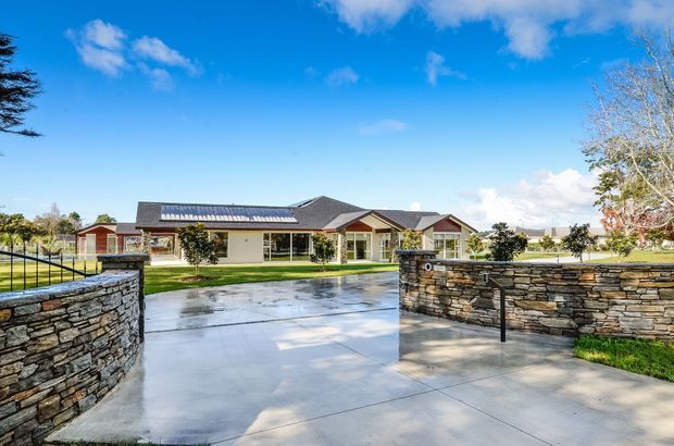 A single-storey suburban home viewed from the front driveway, with a low-pitched dark tiled roof, cream rendered walls, broad glazing, and a covered entry porch. Wet concrete paving leads between curved stacked-stone retaining walls, with manicured lawns and open sky surrounding the property.