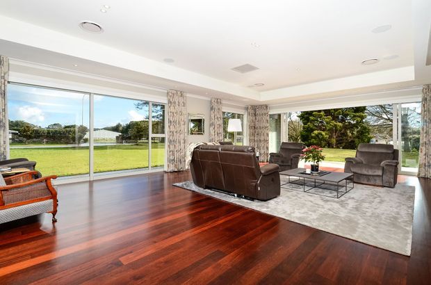 Spacious, bright living room with extensive floor-to-ceiling glazing overlooking a lawn and garden. The room features rich dark timber flooring, a large pale area rug, a brown leather sofa, upholstered armchairs, a glass coffee table, patterned curtains, and a white recessed ceiling with downlights.