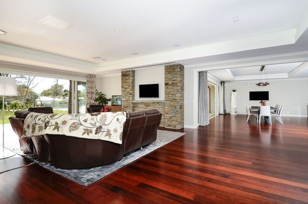 Open-plan living and dining area with rich dark timber flooring, a large brown sectional sofa, stone-clad feature columns framing a wall-mounted television, broad glazing to the garden, and a coffered ceiling. The adjacent dining space continues the polished, spacious interior.