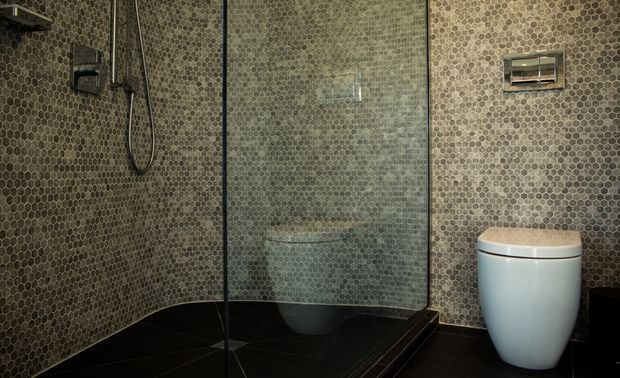 Compact contemporary bathroom with a walk-in glass shower enclosure, wall-mounted shower fixtures, wall-hung toilet, and continuous small hexagonal mosaic tiling in muted grey-brown tones. The floor uses large dark tiles, creating a moody, cohesive finish.