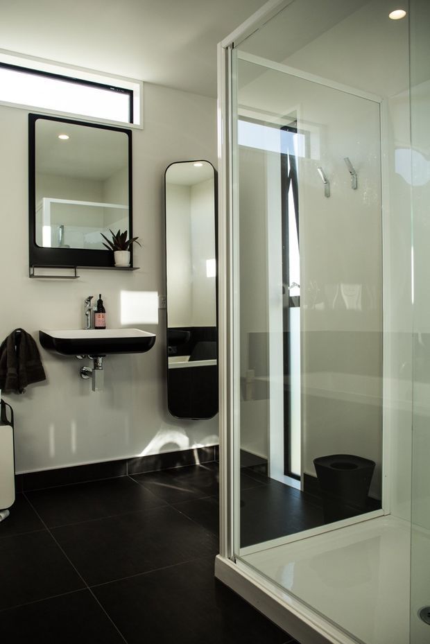 A bright, contemporary bathroom featuring a large glazed walk-in shower enclosure with white framing, a freestanding white bathtub partially visible at the lower right, a wall-mounted black-and-white basin, black-framed mirrors, black floor tiles, white walls, and a tall narrow window. Minimal black fixtures and accessories create a clean monochrome scheme.