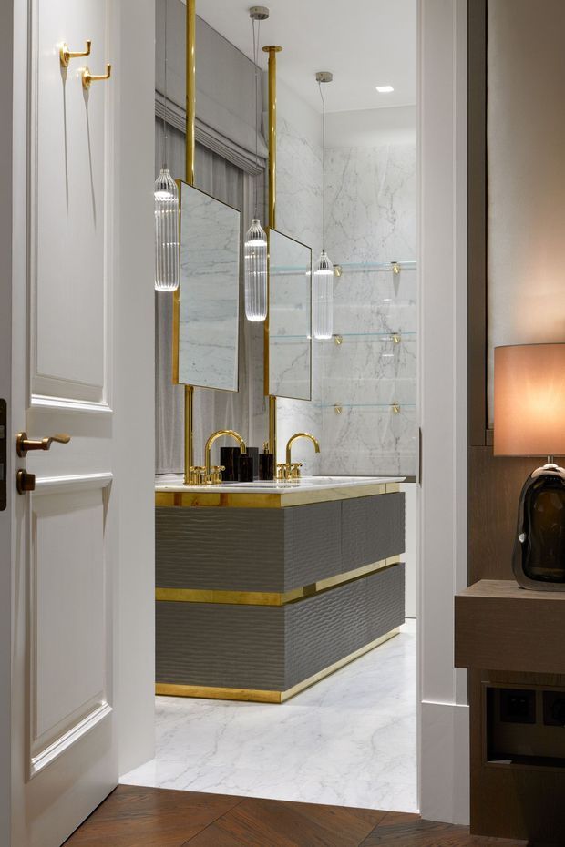 A refined bathroom viewed through an open paneled door, featuring a floating double vanity with textured grey cabinetry, white marble benchtop and wall cladding, polished gold tapware and trim, frameless mirrors, and elegant pendant lights. White marble flooring and glass shelving contribute to the luxurious, highly detailed interior.