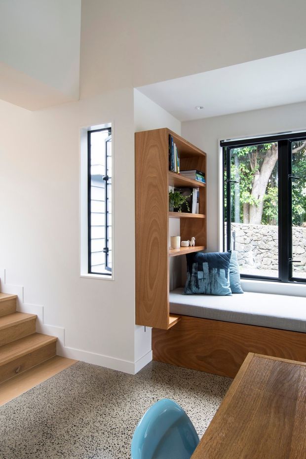 Bright contemporary interior nook combining a staircase, built-in timber shelving and window seat beside black-framed glazing. The space features warm wood joinery, pale walls, terrazzo flooring, a compact desk or table, and views to a stone retaining wall and trees outside.