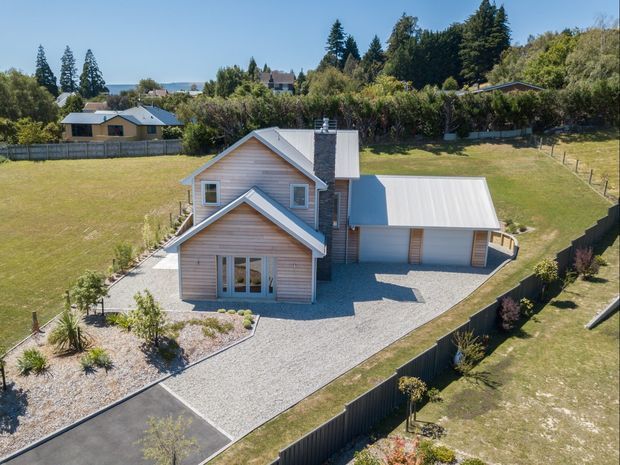 Aerial view of a detached residential house on a landscaped, gently sloping suburban site. The home has pale horizontal weatherboard cladding, steep gabled roof forms with light metal roofing, a prominent central chimney, and an attached garage. Gravel driveways and paths surround the house, with lawns, garden beds, hedges, fencing, and neighbouring homes visible.