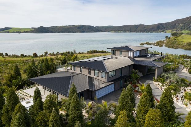 A large contemporary multi-level home with dark cladding, extensive pitched roof forms, and broad glazing, viewed from above within a landscaped garden. The property overlooks a calm coastal inlet or lake, with planted paths, retaining elements, and outdoor terraces surrounding the house.