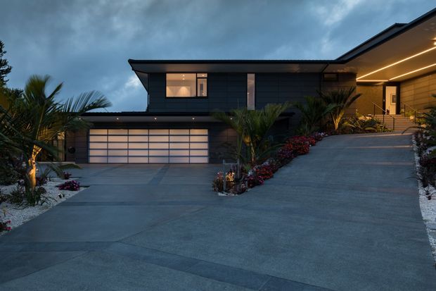 Contemporary two-storey residential facade photographed at dusk, featuring dark horizontal cladding, a wide illuminated garage door, a covered entry with warm linear lighting, broad concrete driveways, and tropical landscaped planting.