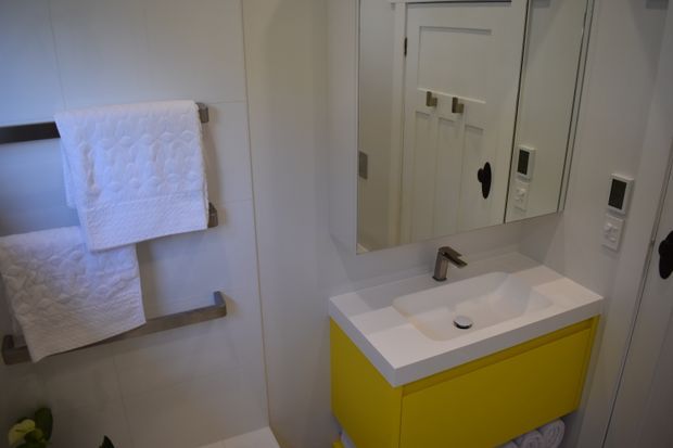 Compact contemporary bathroom featuring a bright yellow floating vanity with an integrated white basin and chrome tapware. White tiled walls, a large mirrored cabinet, and white textured towels on brushed metal rails create a clean, cheerful interior.