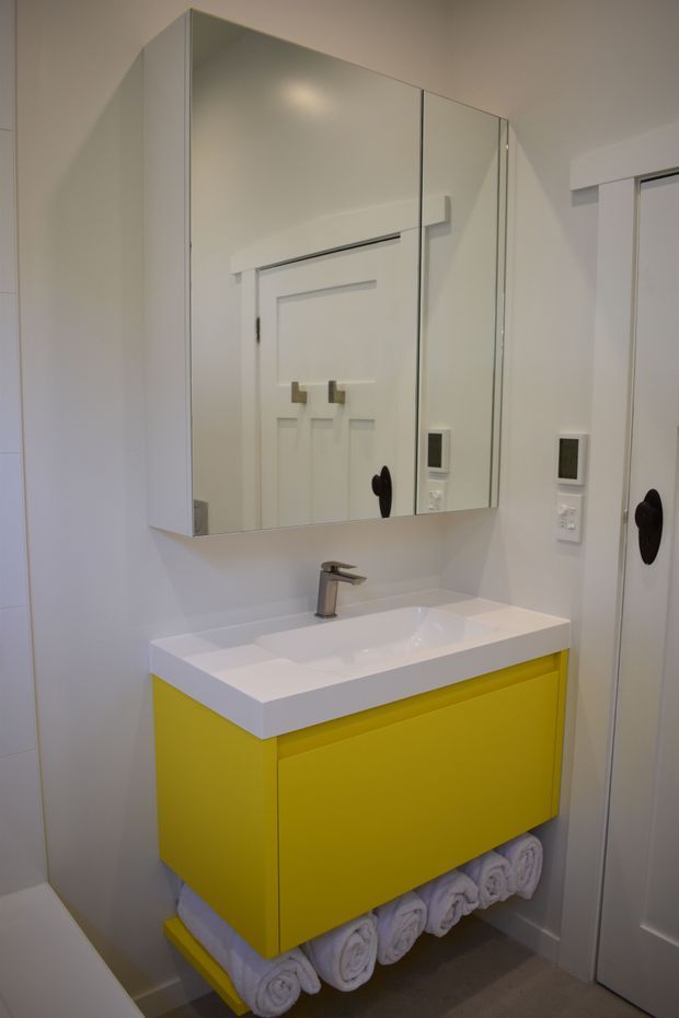 Compact contemporary bathroom vanity with a vivid yellow floating cabinet, white integrated basin and benchtop, chrome mixer tap, and mirrored wall cabinet above. Rolled white towels are stored on an open lower shelf, with a white paneled door and light neutral finishes surrounding the vanity.