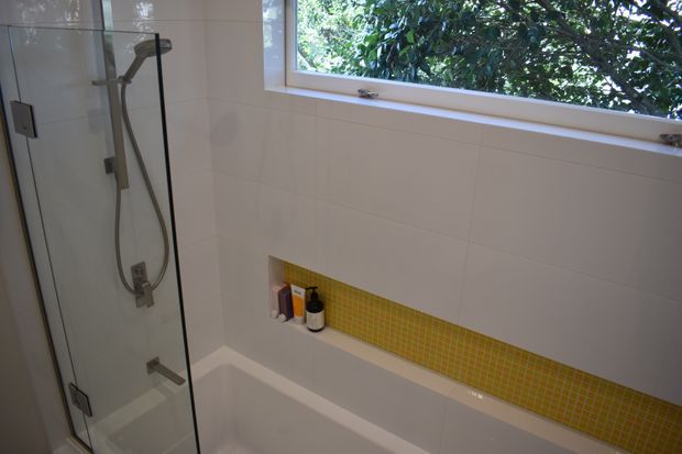 A bright contemporary bathroom featuring a white tiled bath and shower area, frameless glass shower screen, wall-mounted shower fittings, and a narrow yellow mosaic tile feature band with recessed toiletries shelf. A horizontal window brings in natural light and overlooks leafy greenery.