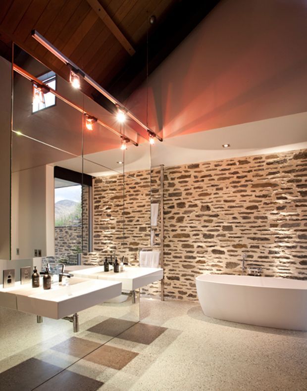 A spacious contemporary bathroom with a freestanding white bathtub, twin floating vanities and large mirrors. The room combines a rustic exposed stone feature wall and polished aggregate-style flooring with a dramatic timber-lined pitched ceiling, exposed dark structural beams, and warm accent lighting. A small high window provides a view toward a mountainous landscape.