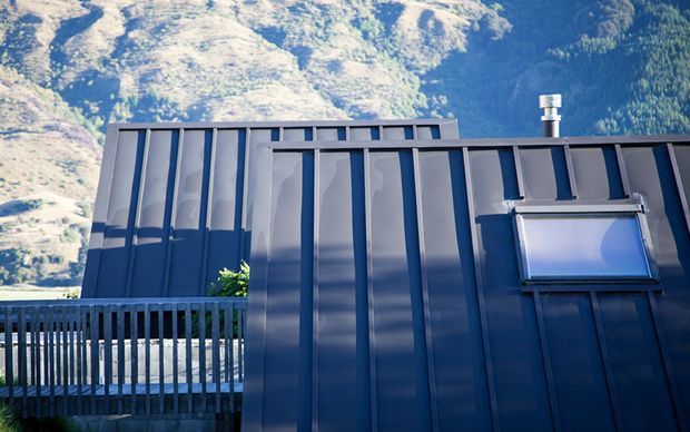 Close exterior view of a dark blue-grey metal-clad building with prominent vertical ribs, a low sloping roof edge, rooftop vent, and a small projecting skylight or window. A timber or metal-railed deck is visible in the foreground, with steep rocky and forested mountain terrain in the background.