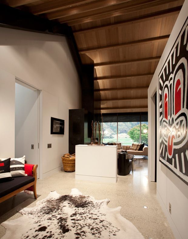 Interior view of a contemporary open-plan living area with a warm timber-lined pitched ceiling, polished light terrazzo or concrete-style flooring, white walls, and bold black, white, and red graphic wall art. A compact black-and-red lounge chair, cowhide rug, kitchen or bar island, and seating area are visible, with large glazing opening toward greenery outside.