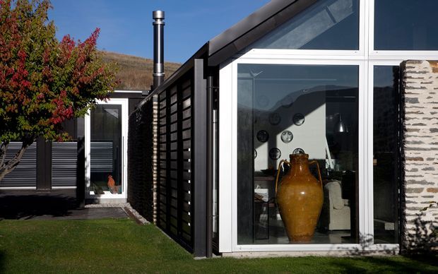 Exterior view of a contemporary single-storey building with black horizontal cladding, large white-framed glazed openings, a sloping roofline, and a textured stacked-stone feature wall. A dark timber privacy screen and landscaped lawn define the approach, while interior furnishings and a large ceramic vessel are visible through the glazing.