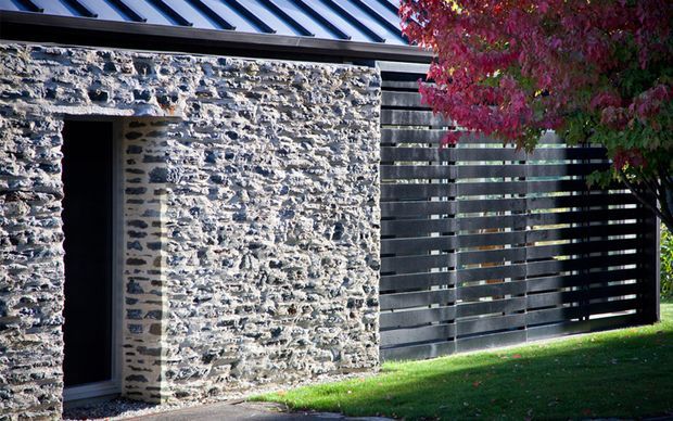 Exterior view of a contemporary structure featuring rough natural stone masonry walls, a dark standing-seam metal roof, and a horizontal dark timber or metal slatted screen. A narrow doorway opens into a shadowed interior, with landscaped lawn and red foliage alongside.