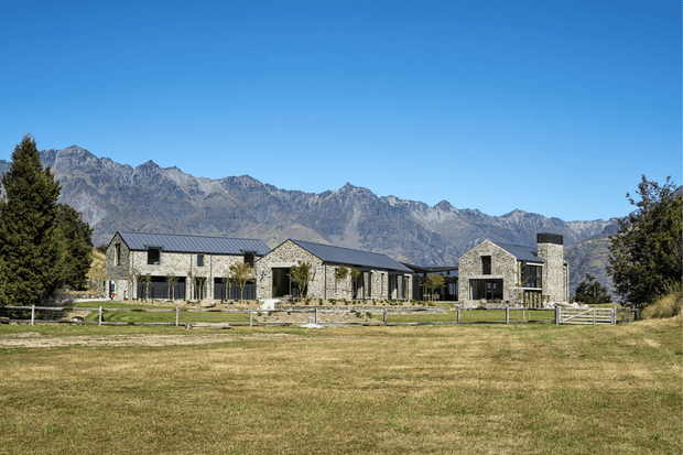 A large stone-clad rural residence set behind an open lawn, composed of several connected gabled volumes with dark pitched metal roofs, tall stone chimneys, and expansive mountain views in the background. The property has a formal driveway or lawn frontage, fencing, mature trees, and a spacious alpine setting.