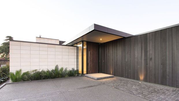 A modern residential entrance viewed from the driveway, featuring a flat-roofed projecting canopy, dark vertical timber cladding, a large timber entry door, pale rectangular masonry or rendered wall panels, and warm recessed and uplighting. Low architectural planting borders the paved approach.